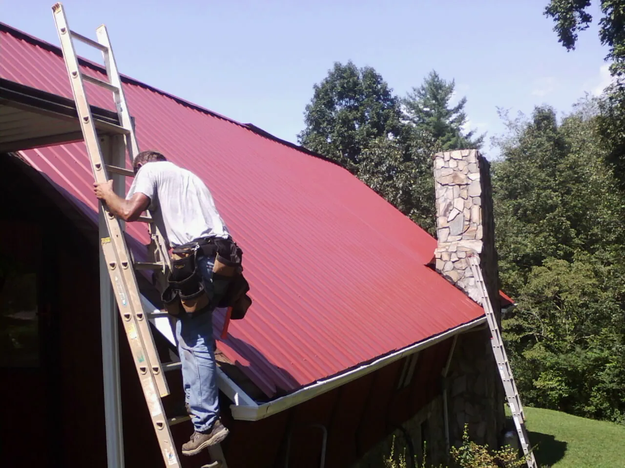 Roofer on a ladder beside a red metal roof and stone chimney — Soffit & Fascia Repair by Roofing Brothers in Mercer Island